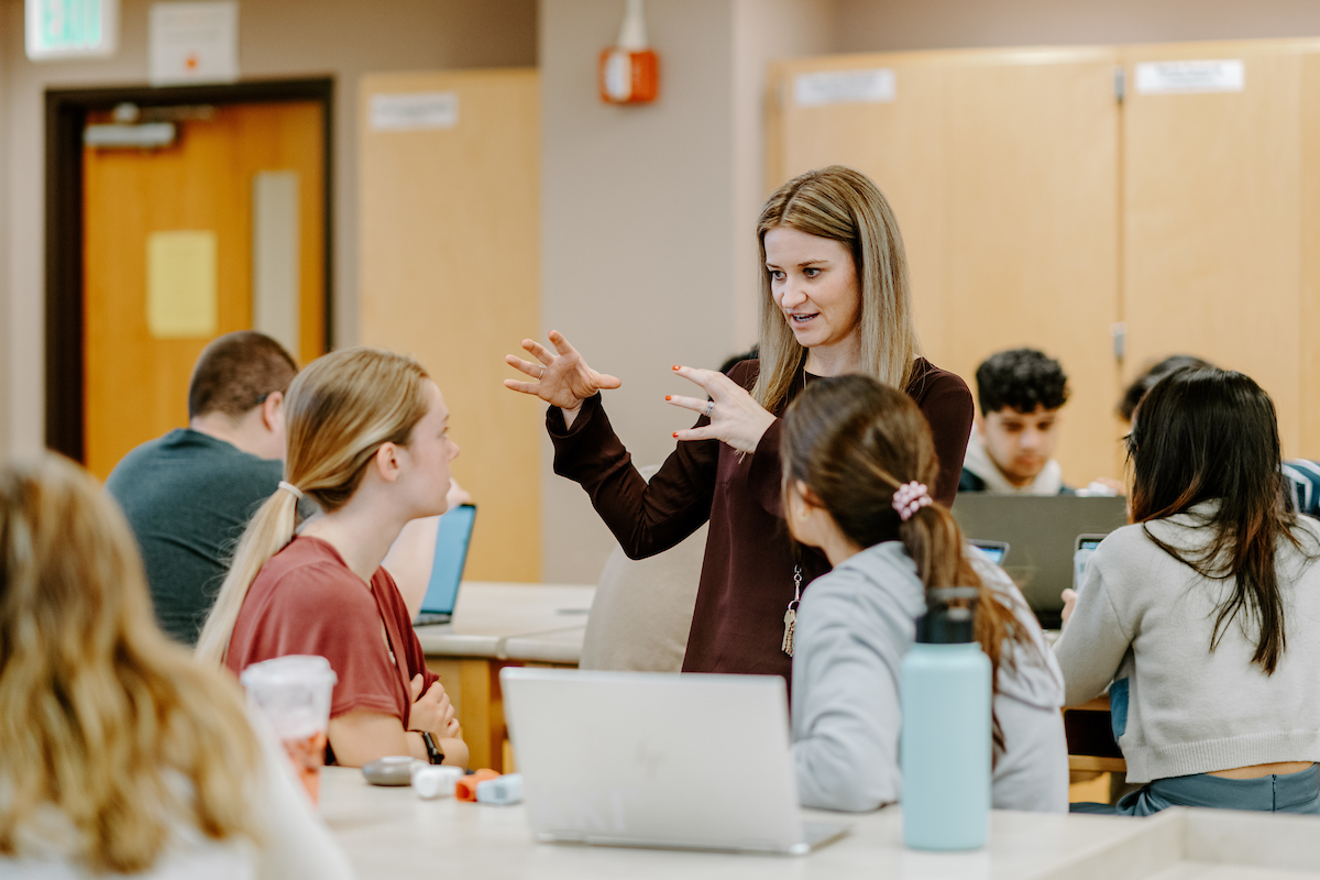 pictured: students sit in a classroom around a laptop, looking back at their professor